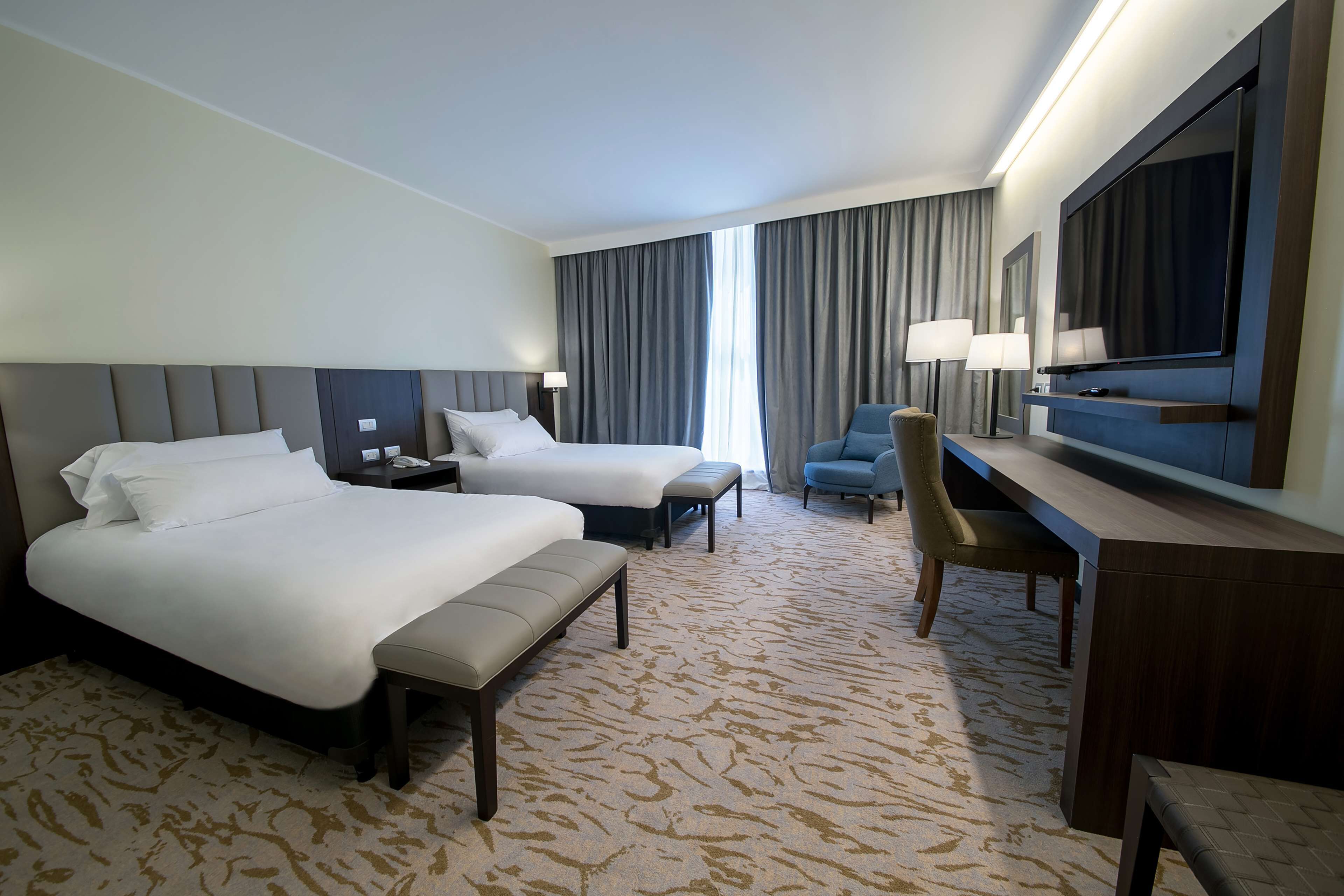A hotel room with two beds, a desk, a chair, a TV, and a large window with curtains.