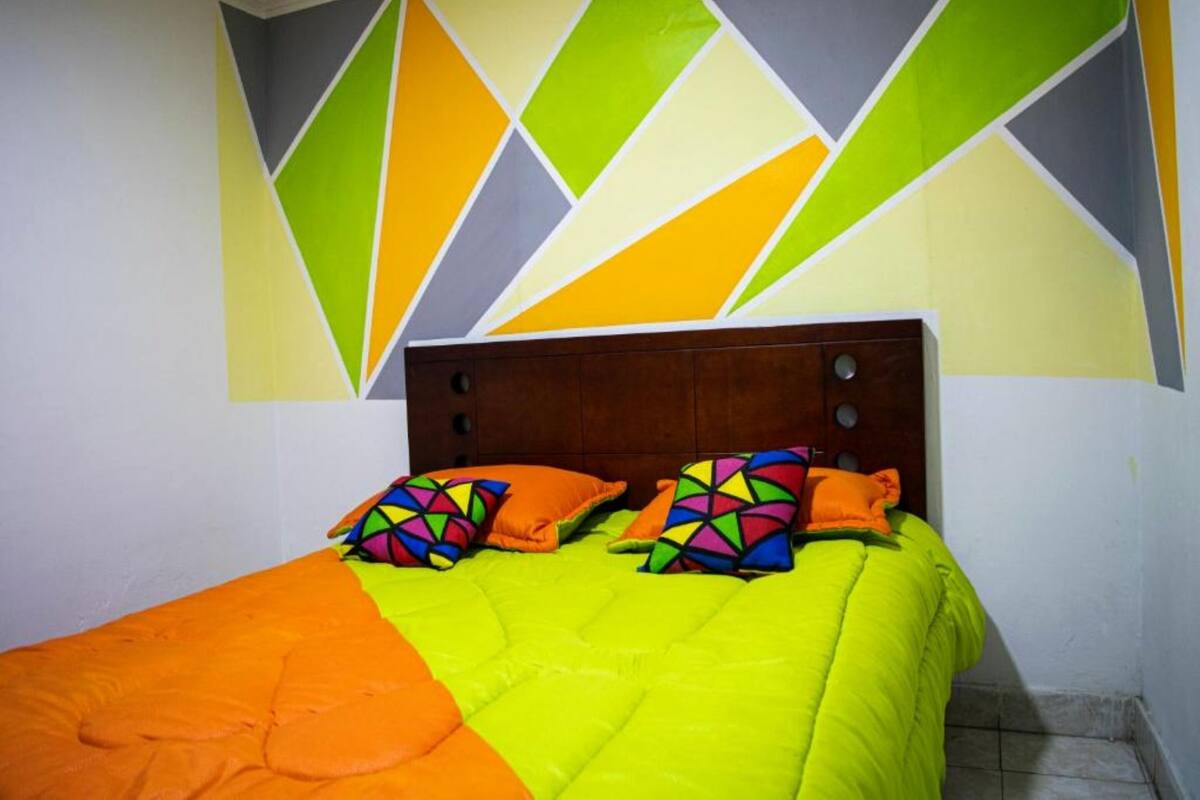 Ein Bett mit einer leuchtend orangefarbenen und grünen Tagesdecke, einem Holzkopfende und farbenfrohen, geometrisch gemusterten Kissen.