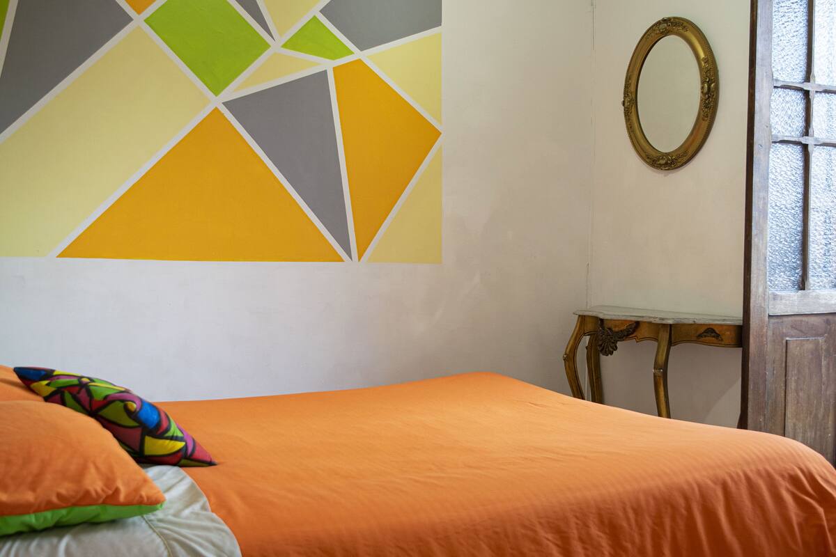 Ein Zimmer mit einem Bett, farbenfrohen Kissen, einer geometrischen Wanddekoration, einem Spiegel und einem Fenster mit Holzrahmen.