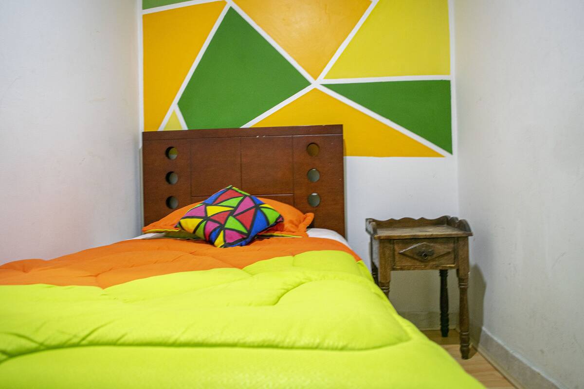 Ein Bett mit hölzernem Kopfteil, einem farbenfrohen geometrischen Kissen und einer orange-grünen Decke.