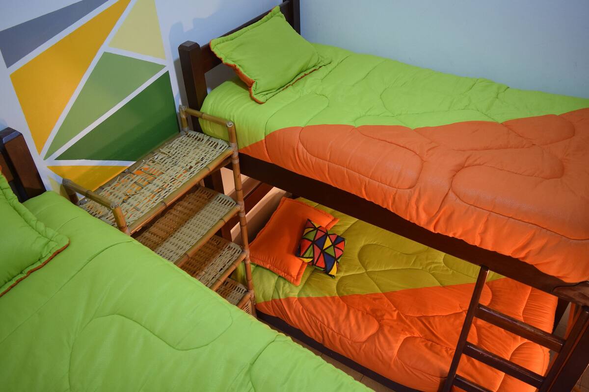 Ein Stockbett mit grün-orange gestreuter Bettwäsche, ein gewebter Stuhl und ein geometrisches Wandbild.