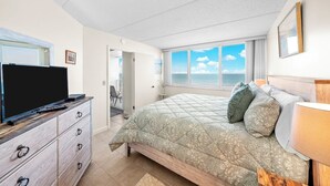 A bedroom with a bed, a TV, a dresser, and a window with a view of the sea.