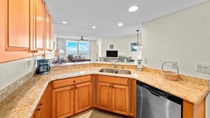 A kitchen with wooden cabinets, granite countertops, a stainless steel dishwasher, and a large window overlooking a balcony.