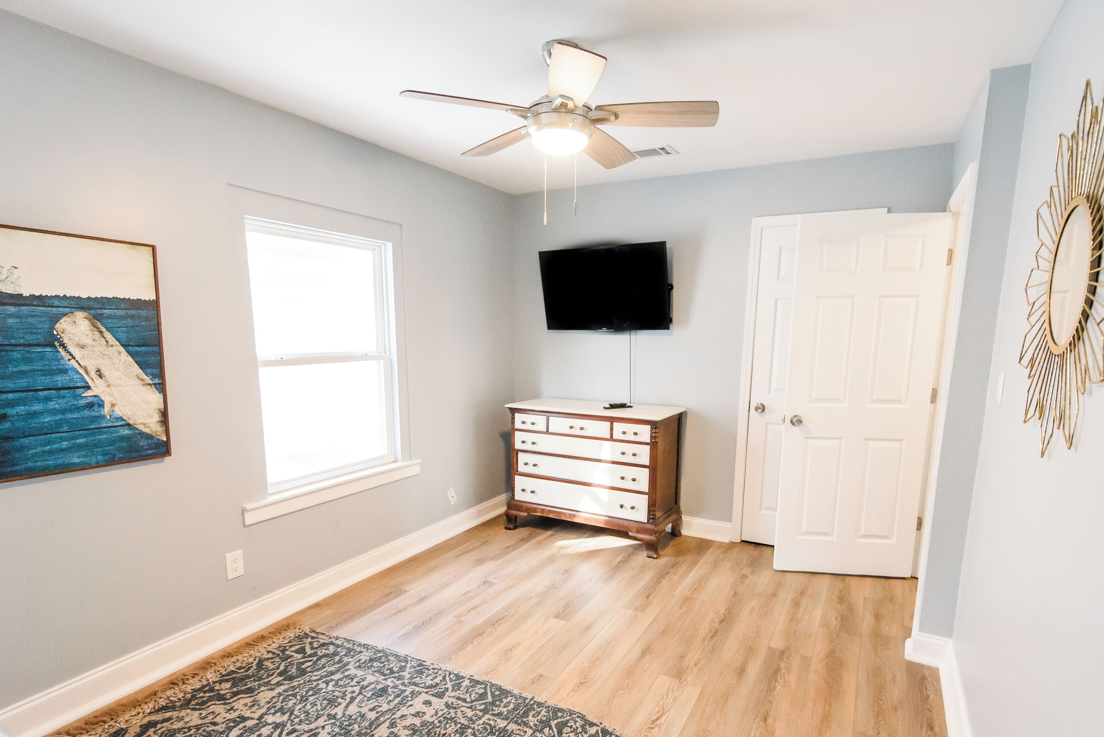 A room with hardwood flooring, a ceiling fan, a flat-screen TV mounted on the wall, a wooden dresser, and a patterned rug.