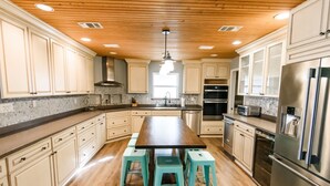 A modern kitchen with wooden ceiling, white cabinets, a central island with stools, stainless steel appliances, and a large window providing natural light. - Lakefront Home Close to the Beach! Sleeps 16 (Santa Rosa Beach)