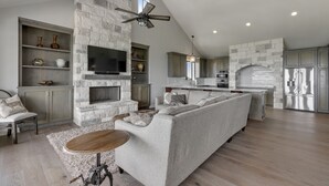 A spacious living area with a stone fireplace, a large gray sofa, a wooden coffee table, and a TV mounted on the wall. - “WATERS-EDGE” A WATERFRONT home! Hot Tub!! VIEWS!!! (Canyon Lake)