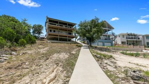 A multi-story building with a concrete pathway leading to it, surrounded by trees and a clear sky. - “WATERS-EDGE” A WATERFRONT home! Hot Tub!! VIEWS!!! (Canyon Lake)