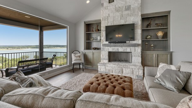 A modern living room with a stone fireplace, a flat-screen TV, and a large window overlooking a scenic view. - “WATERS-EDGE” A WATERFRONT home! Hot Tub!! VIEWS!!! (Canyon Lake)
