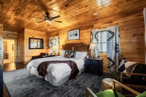 Un dormitorio de cabaña de madera con cama, mesitas de noche, una silla y un ventilador de techo.