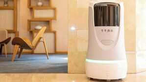 Ein weißer, tragbarer Heizlüfter mit digitaler Anzeige und Einschalttaste, platziert auf einem Fliesenboden in der Nähe eines Holzstuhls und eines Regals mit Dekoration.