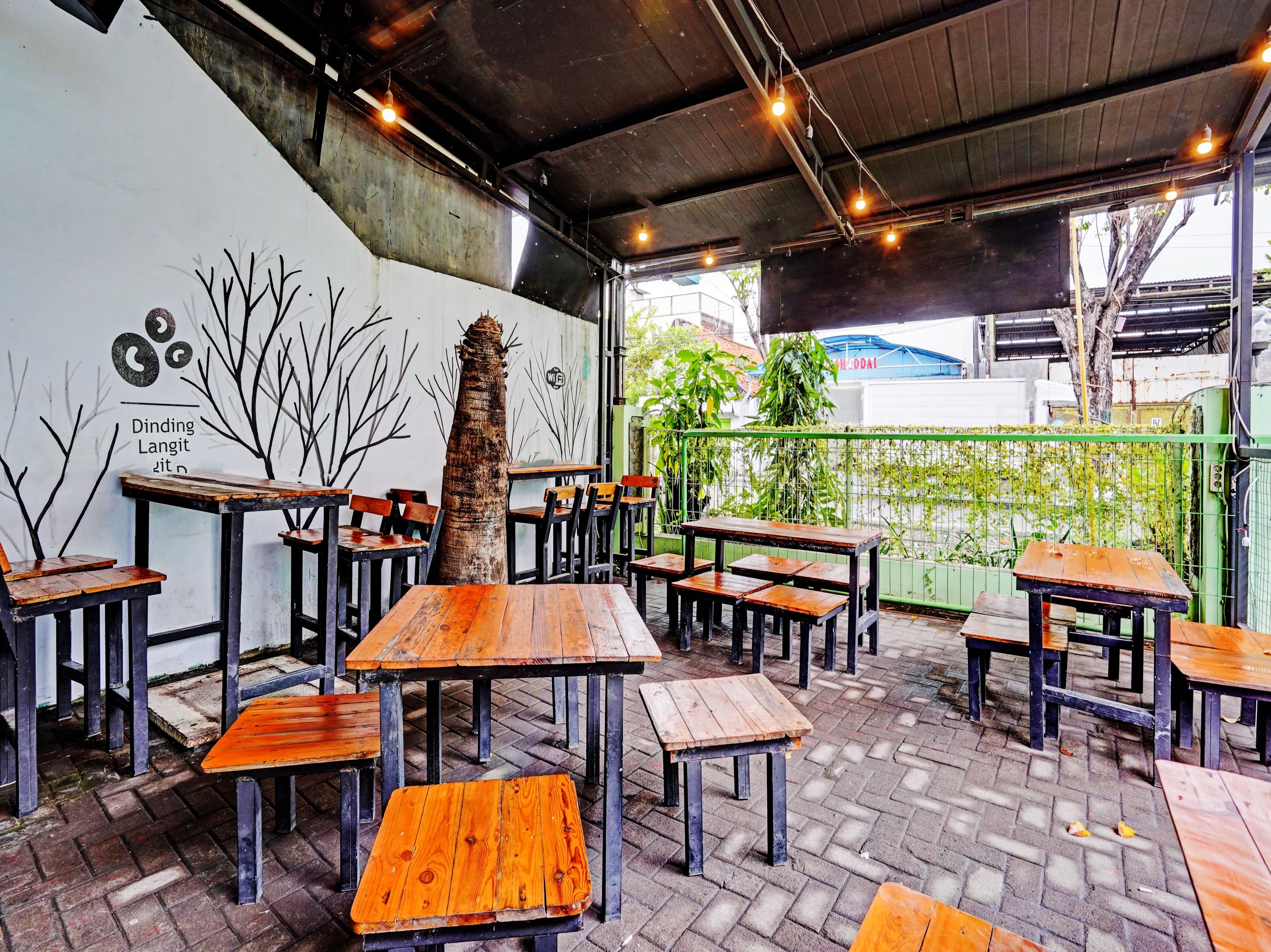 An outdoor seating area with wooden tables and benches, a mural on the wall, and a green fence in the background.