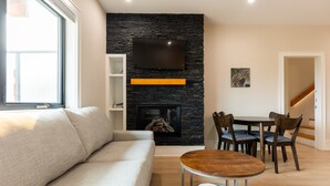 A modern living room with a stone fireplace, a flat-screen TV mounted above it, a sofa, a dining table with chairs, and a staircase in the background. - Luxury. Steps to the Timberline Chairlift (Fernie)