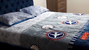 A bed with a blue headboard, a grey blanket with nautical symbols, and a wooden nightstand with a folded blanket.