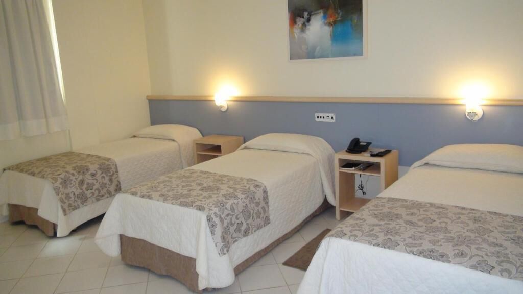 A hotel room with three single beds, each with a bedside table and a lamp.