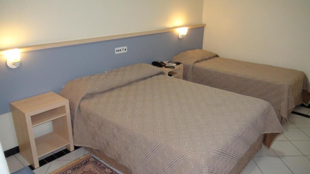 Una habitación con dos camas, cada una con cabecera y mesitas de noche. Hay un enchufe en la pared y una pequeña estantería.
