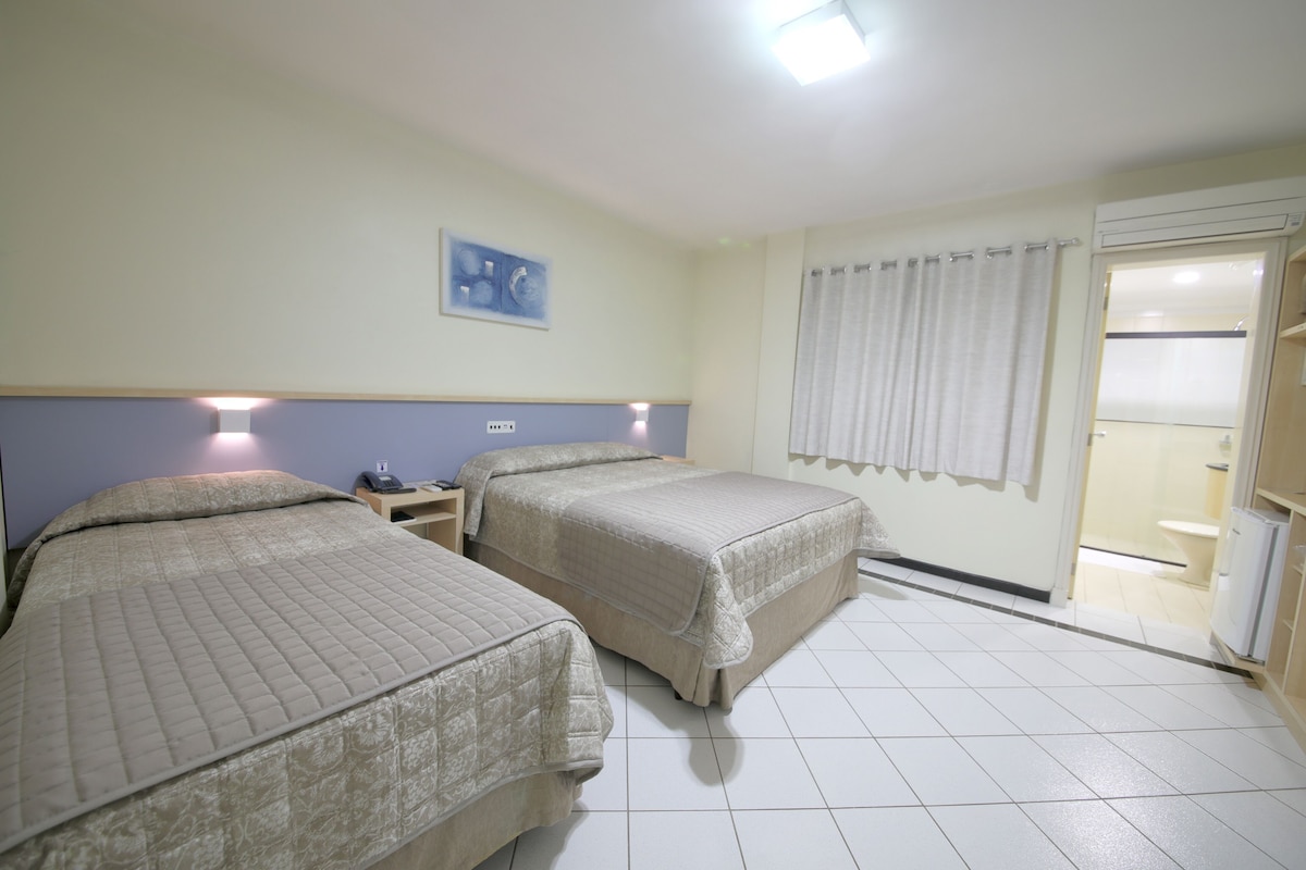 A hotel room with two beds, a nightstand, a wall-mounted phone, a painting, a curtain, and a bathroom visible through an open door.