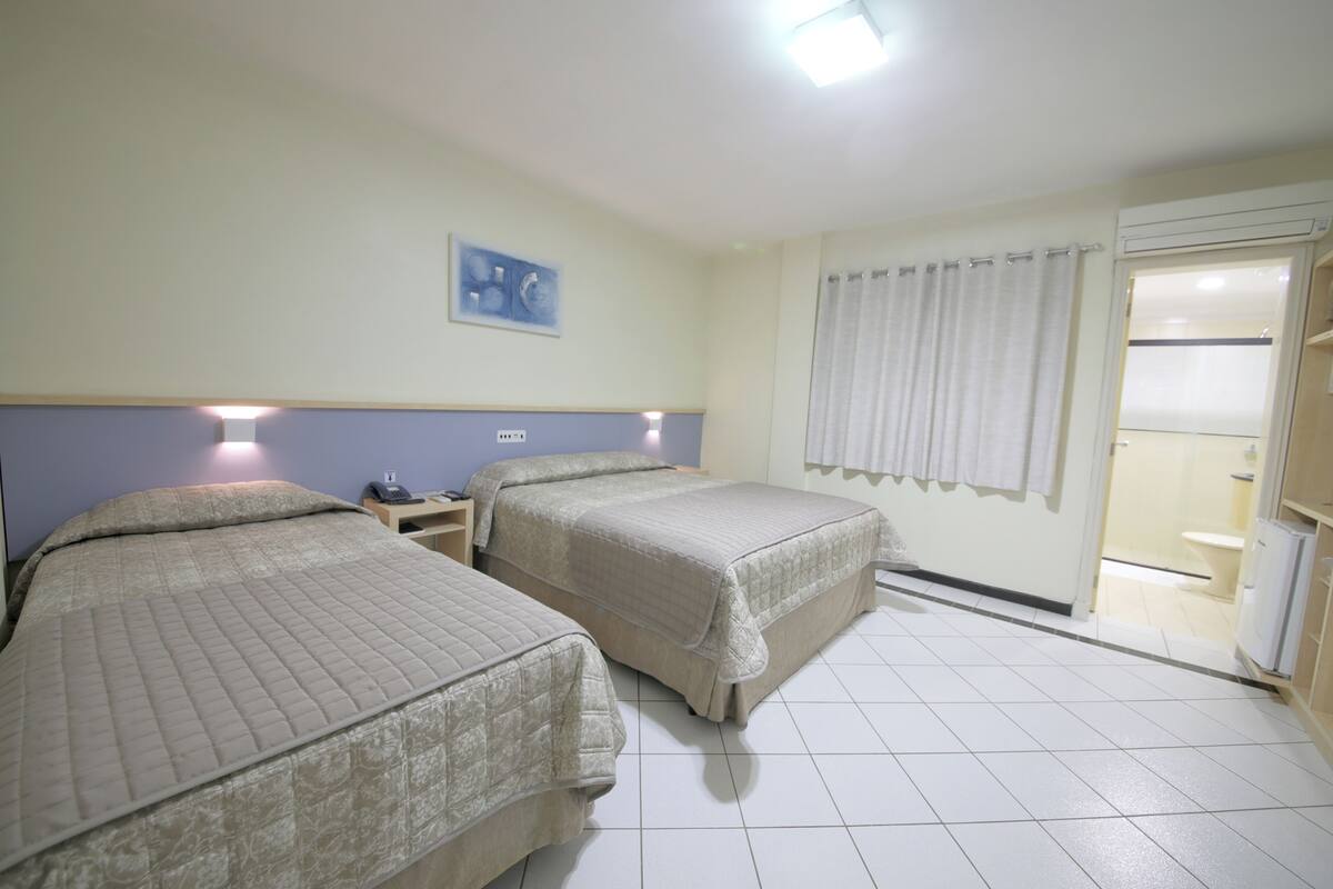 A hotel room with two beds, a nightstand, a wall-mounted phone, a painting, and a bathroom visible through a door.