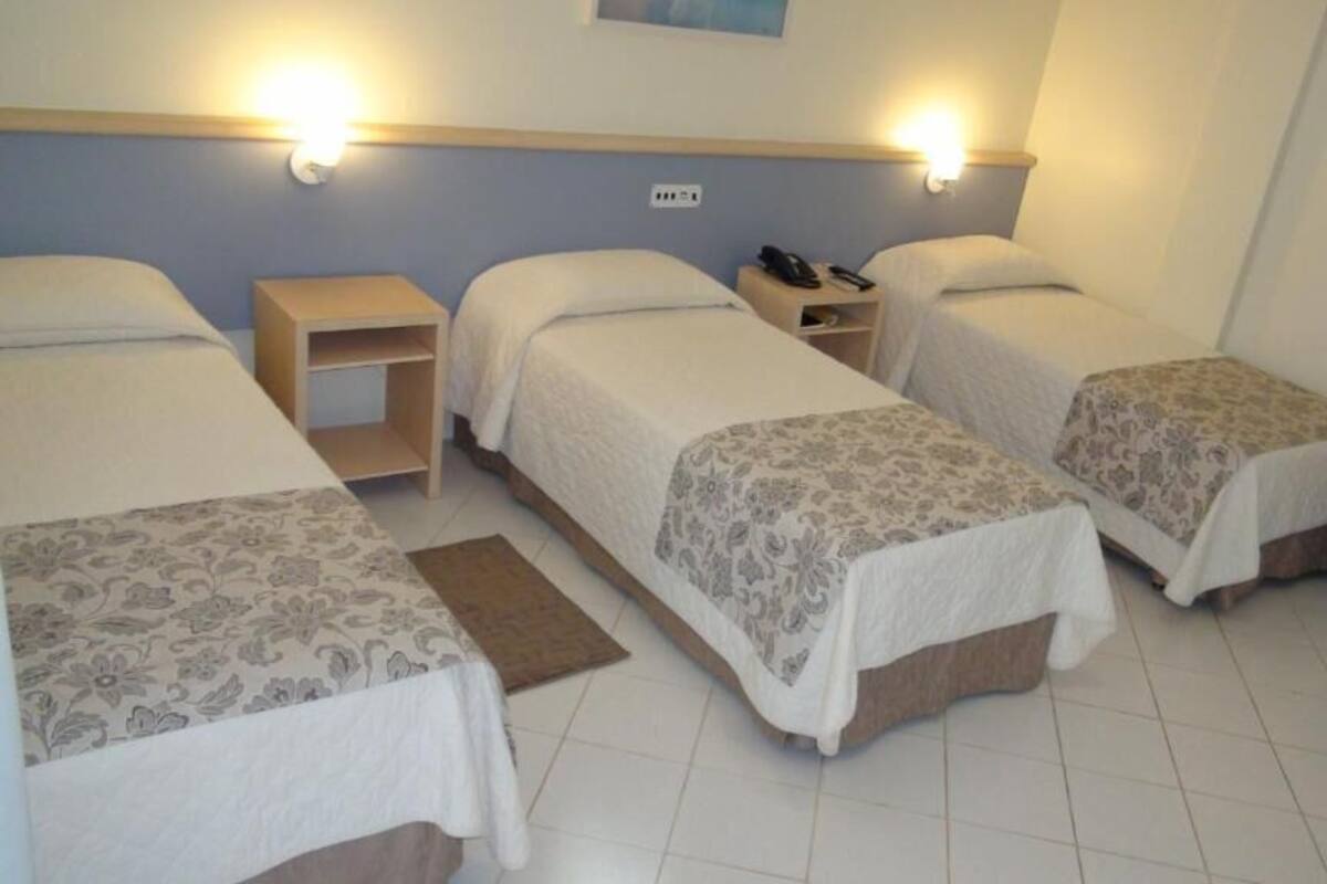 A hotel room with three single beds, each with a bedside table and lamp.