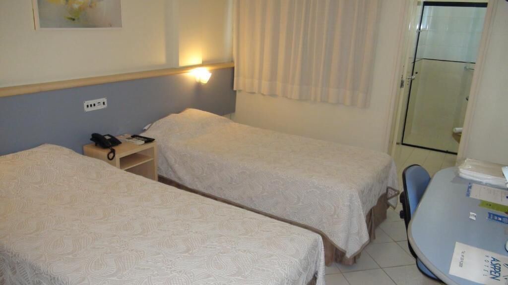 Une chambre d’hôtel avec deux lits simples, une table de chevet avec un téléphone et des documents, un petit bureau avec une chaise, et une salle de bain visible à travers une porte en verre.