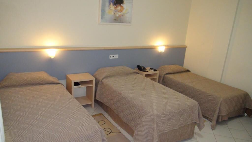 Quarto com três camas de solteiro, cada uma com cabeceira e mesa de cabeceira. Há uma luminária fixada na parede sobre as camas e uma pequena prateleira entre elas.
