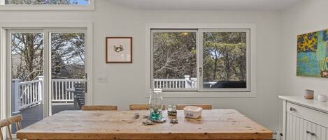 Un coin repas avec une table en bois, des chaises et un banc. Il y a des fenêtres donnant sur des arbres et une terrasse à l’extérieur.