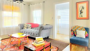 Una sala de estar con un sofá gris, un sillón azul, una mesa de centro de madera y un tapete colorido. Hay un baúl rojo con libros encima y una lámpara en la esquina.