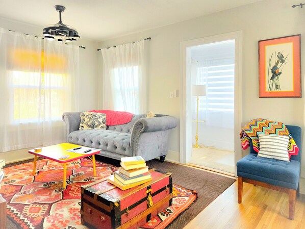 A living room with a grey sofa, a blue armchair, a wooden coffee table, and a colorful rug. There is a red trunk with books on top, and a lamp in the corner.