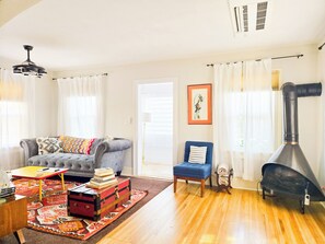 A living room with a sofa, a blue armchair, a small table with books, a colorful rug, and a black stove.