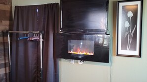 A room with a flat-screen TV mounted on the wall, a small fireplace below it, and a framed picture of flowers. - Cozy Tiny Cabin with a small porch and firepit. (Kempner)