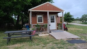 A small, rustic cabin with a porch, a welcome sign, and a picnic table. - Cozy Tiny Cabin with a small porch and firepit. (Kempner)