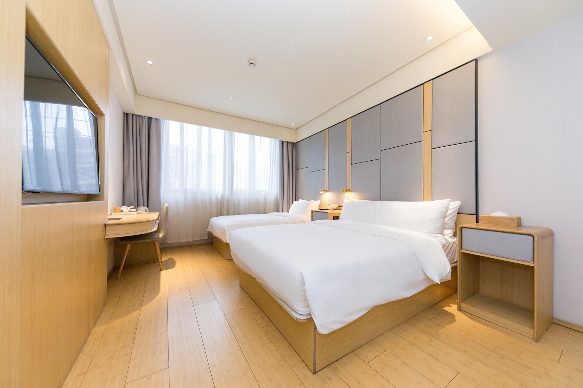 A modern hotel room with a large bed, bedside tables, a desk with a chair, and a large window with curtains.