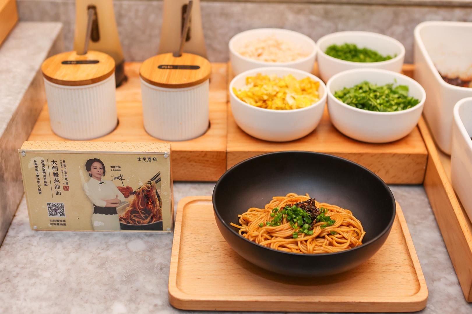 Una ciotola di noodles con condimenti su un tagliere di legno, circondata da ciotole di ingredienti e una scatola con il menu.