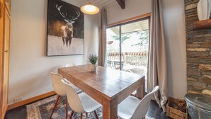 Uma área de refeições com mesa de madeira, cadeiras brancas, pintura de um veado e parede de pedra.
