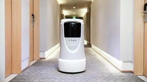 A white, modern, automated room service robot in a hotel corridor with wooden doors and a tiled floor. - Ji Hotel (Shanghai)