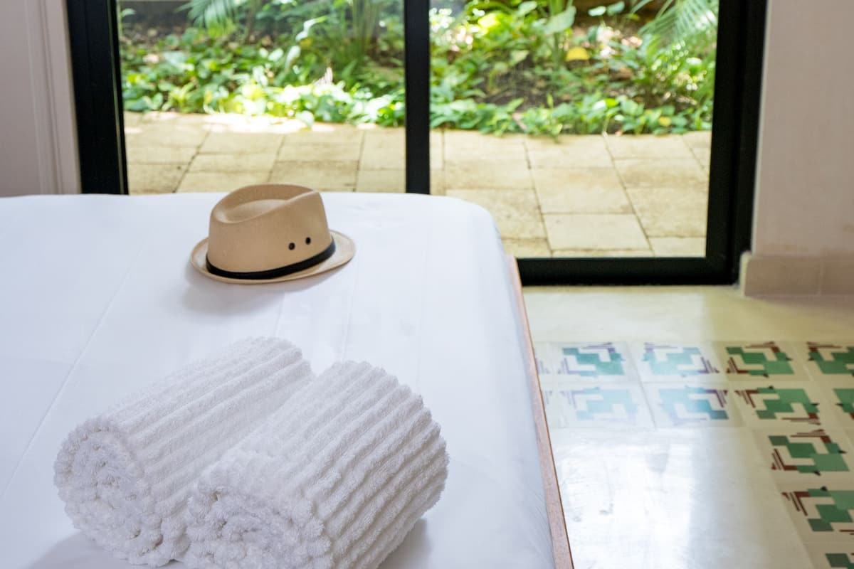 A bed with white linens and a hat on it, a hammock in the background, and a view of a garden through a glass door.