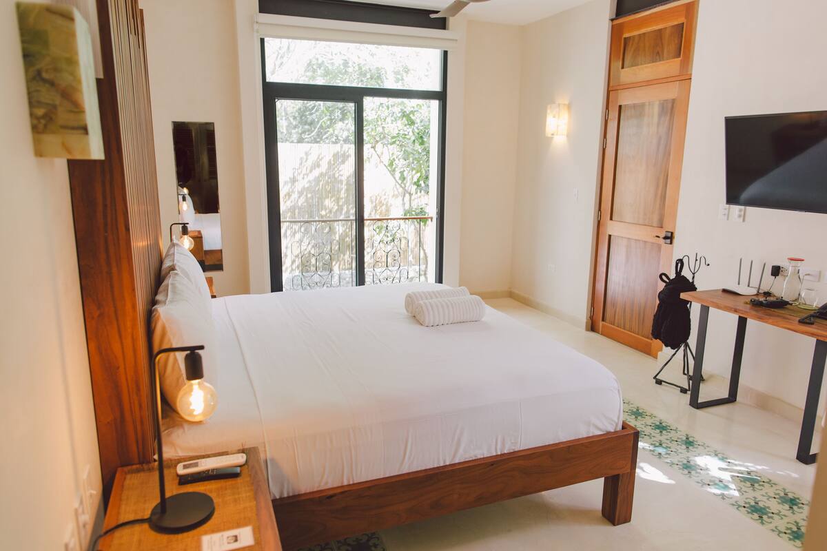 A bedroom with a large bed, a bedside table with a lamp, a TV on the wall, and a window with a view of greenery.