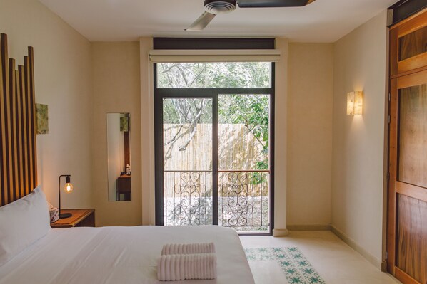A hotel room with a bed, a nightstand, a mirror, a window with a view of greenery, and a door leading to an outdoor area.