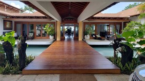 A wooden pathway leading to a covered area with a pool, surrounded by tropical plants and a car.