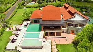 A large house with a swimming pool, surrounded by lush greenery and a road.