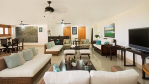 A spacious living room with a large sofa, two armchairs, a coffee table, and a TV. There are ceiling fans, a stone wall, and a dining area with a table and chairs.