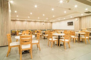 A spacious dining area with round tables and wooden chairs, featuring a clock on the wall and recessed lighting.