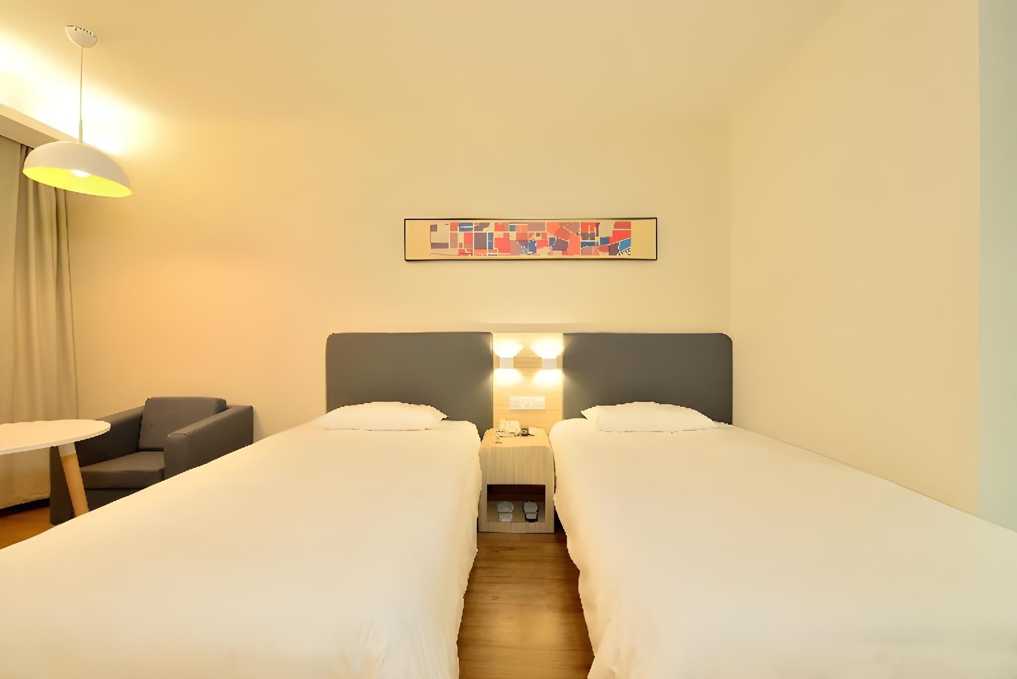 Habitación de hotel con dos camas individuales, una mesita pequeña, una silla, un escritorio con lámpara y una obra de arte colgada en la pared.