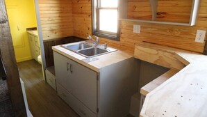 A compact kitchen with wooden walls, a sink, and a window. - Secluded Wooded Charmant Chalet  "Theme"  Tiny Home - Off Grid (Aylette)
