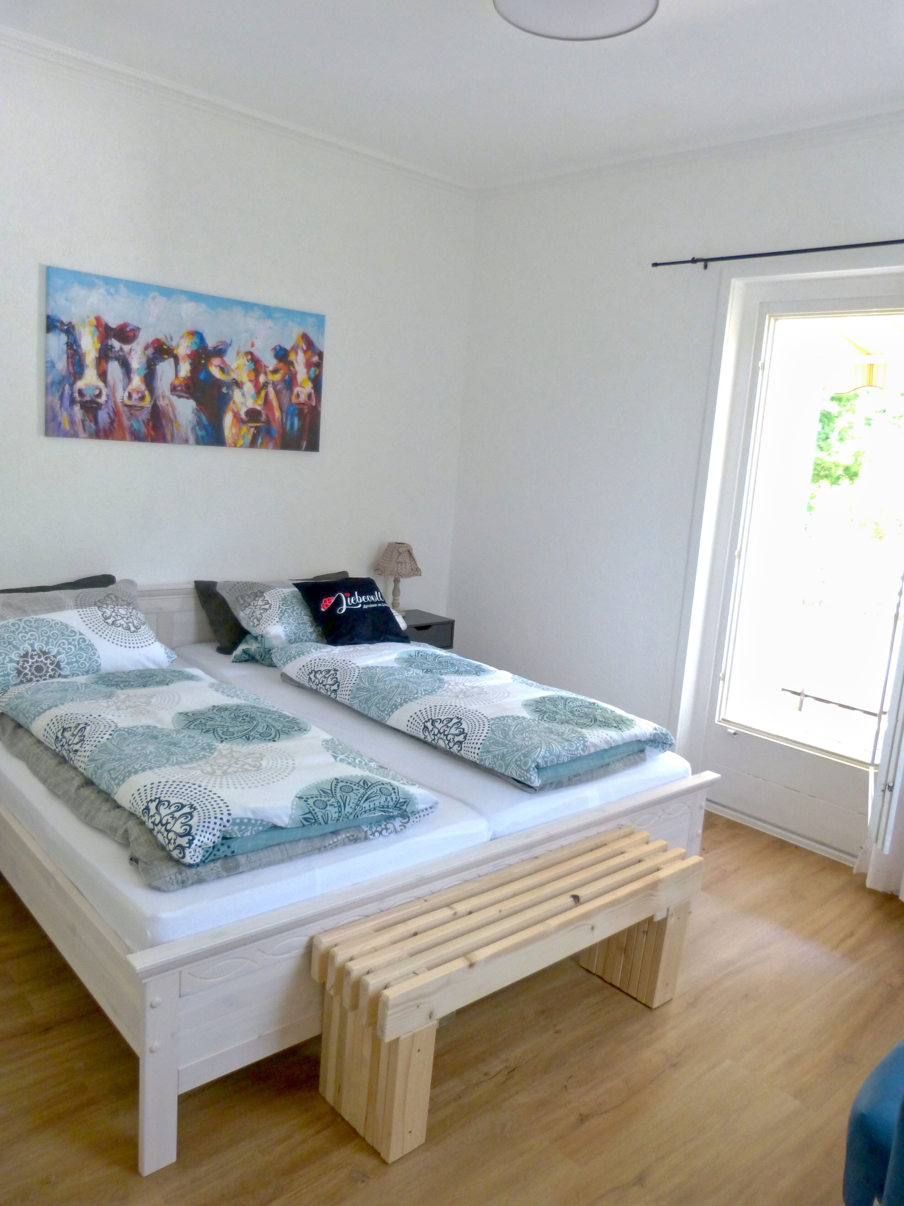 A bedroom with a bed, pillows, a bench, a painting, and a window.