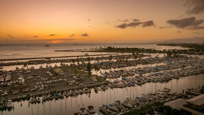 Uma marina com vários barcos atracados, uma área de estacionamento com carros e vista para o pôr do sol sobre a água.