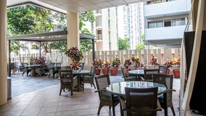 An outdoor patio area with round tables and wicker chairs, surrounded by potted plants and a white fence. - Aqua Aloha Surf Waikiki 313 | Downtown City Views – Pool & BBQ (Honolulu)