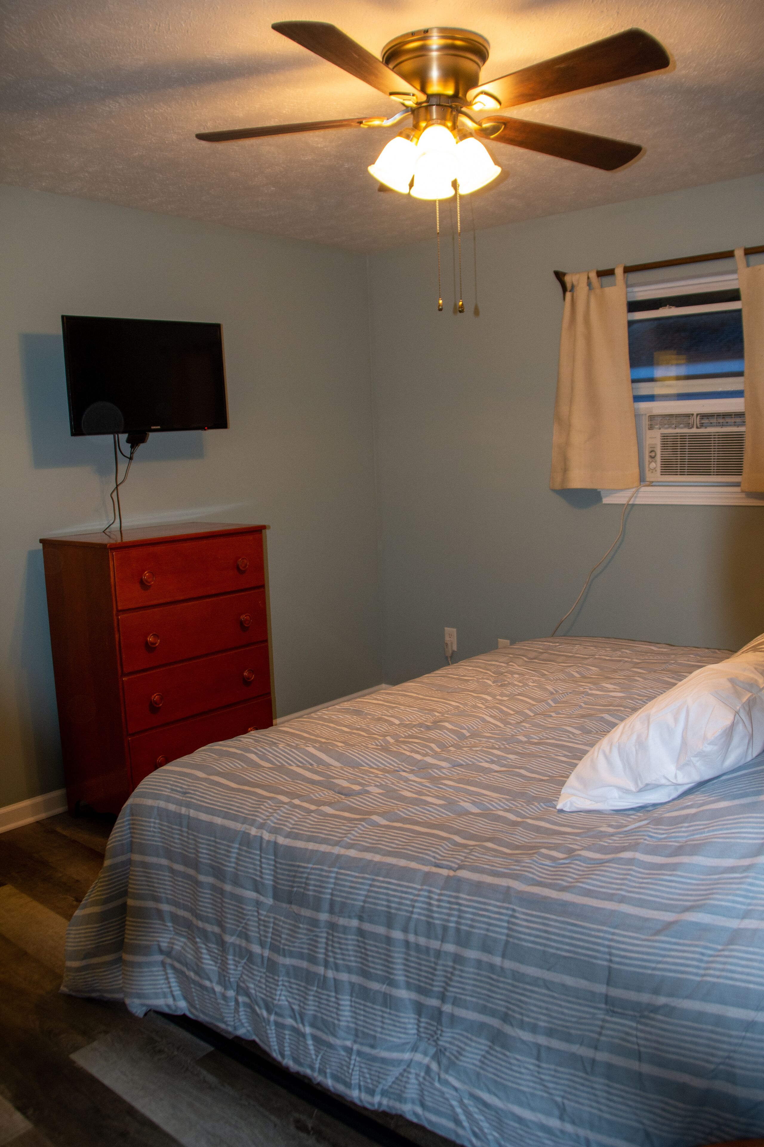 Une chambre à coucher comprenant un lit, un ventilateur de plafond, une commode, un téléviseur et une fenêtre avec des rideaux.