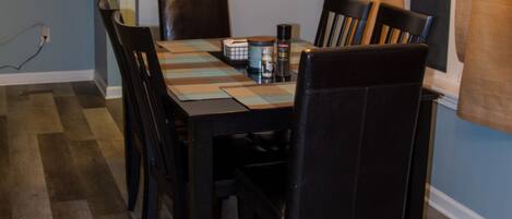 A dining area with a table set for six, surrounded by six chairs. A chandelier hangs from the ceiling, and a window with curtains is visible to the right.