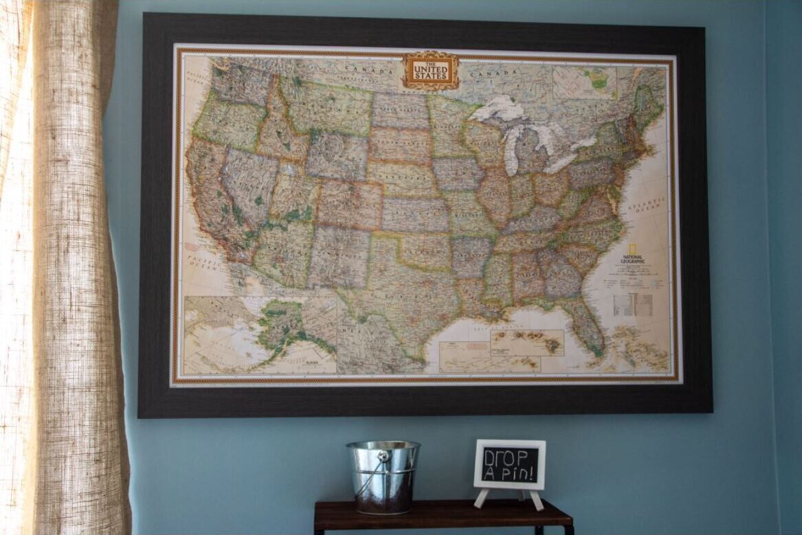 Une carte encadrée des États-Unis est accrochée au mur, avec un seau et une pancarte sur laquelle on peut lire : « DÉPOSEZ UNE ÉPINGLE ! »