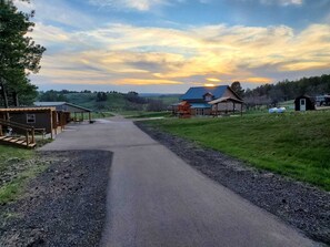 Une route de campagne mène à une grande maison avec un toit bleu, entourée de champs verdoyants et d’une petite construction en bois située sur la gauche.